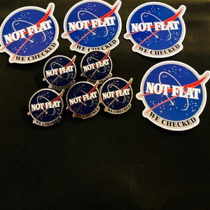 May include: A set of eight stickers and pins featuring a blue and red graphic of a rocket ship with the text "NOT FLAT WE CHECKED".