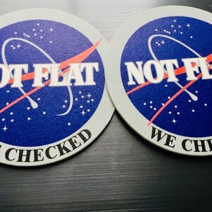 May include: Two round, white coasters with a blue and red NASA-inspired design. The text "NOT FLAT WE CHECKED" is printed on each coaster.