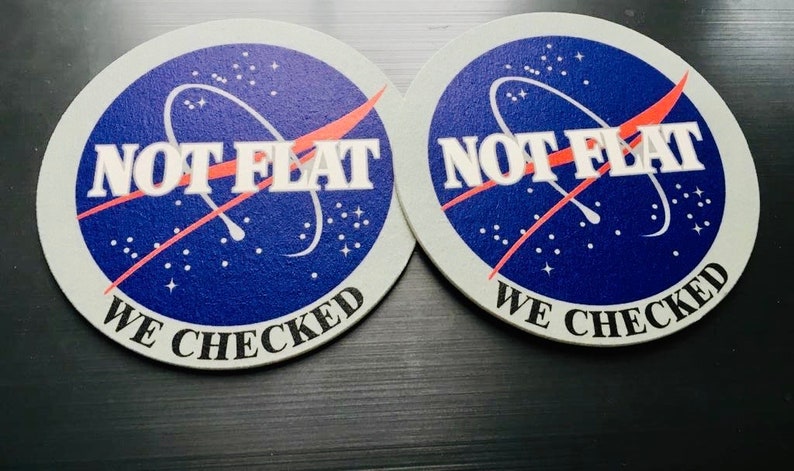 May include: Two round, white coasters with a blue and red NASA-inspired design. The text "NOT FLAT WE CHECKED" is printed on each coaster.