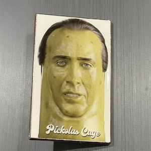 May include: A rectangular, off-white box featuring a portrait of Nicolas Cage in a yellow-toned style. The text "Pickolas Cage" is written in a cursive font at the bottom of the image. The box has a brown border.