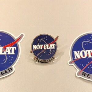 May include: Three stickers with a blue and white NASA-style logo that says "NOT FLAT WE CHECKED" in white letters. The stickers are in different sizes and one is a pin.