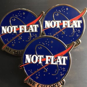May include: Three gold-colored pins with a blue and red NASA-inspired design. Each pin features the text "NOT FLAT" and "WE CHECKED" in white lettering.