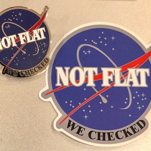 May include: A blue and white sticker with a red and white design. The sticker features a stylized image of a planet with a red and white orbit around it. The text "NOT FLAT WE CHECKED" is printed in white on the sticker.