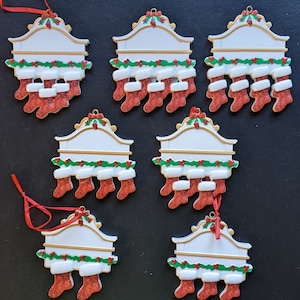 May include: Seven Christmas stocking ornaments. Each ornament has a white sign with gold trim, a garland of green leaves and red berries, and red glitter stockings. A red ribbon is attached for hanging.