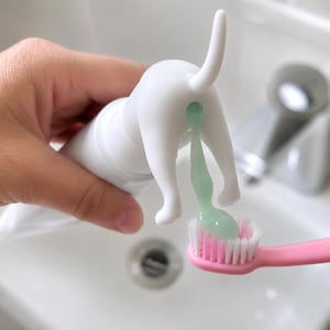 May include: A white plastic toothpaste dispenser shaped like the rear of a dog. The dispenser is being squeezed to release a stream of light green toothpaste onto the bristles of a pink toothbrush. The design features a tail and four legs.