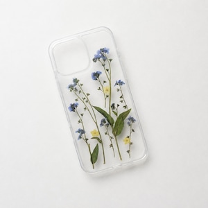 May include: A clear phone case featuring an arrangement of pressed blue forget-me-not flowers and small yellow blossoms with green stems and leaves. The case has a rectangular cutout for a camera lens at the top left corner and a smooth finish.