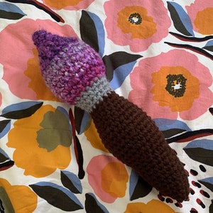 Könnte beinhalten: Ein gehäkeltes Plüschtier in Form eines Pinsels. Der Pinsel ist lila, grau und braun. Das Spielzeug liegt auf einem weißen Stoff mit Blumenmuster.