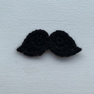 Puede incluir: Un bigote de ganchillo negro. El bigote está hecho de hilo y tiene una textura peluda.