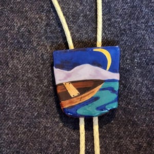 May include: A ceramic bolo tie featuring a blue, green, and purple landscape with a brown boat and a yellow crescent moon.