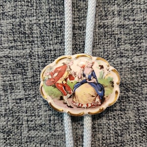 May include: A white porcelain bolo tie featuring a painted scene of a couple in 18th-century attire. The man is wearing a red jacket and the woman is wearing a blue dress. The bolo tie is on a light blue cord.