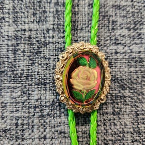 May include: A green bolo tie with a gold oval pendant featuring a yellow rose with green leaves on a pink background.