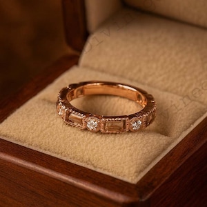 May include: Rose gold eternity ring with alternating baguette and round cut diamonds. The ring is displayed in an open wooden jewelry box with a beige velvet interior. The box has decorative gold accents.