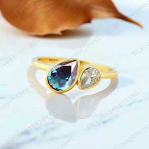Pode incluir: Um anel de ouro com duas pedras preciosas em forma de pêra. Uma é de cor verde-azulado profundo, a outra é transparente. O anel tem um design simples e elegante, com as pedras preciosas lado a lado. A palavra "Divine" é visível no anel.