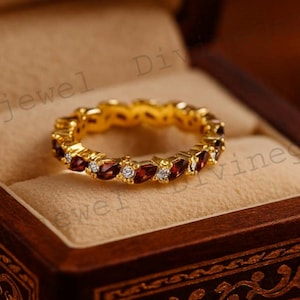 May include: A gold-colored ring with alternating red and white gemstones, displayed inside a wooden jewelry box. The ring features a continuous band of oval-shaped red stones and smaller, round white stones.