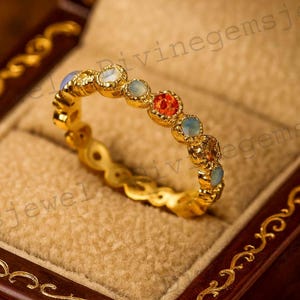 Può includere: Un anello d'oro con una serie di pietre preziose colorate, tra cui pietre blu, arancioni e bianche. L'anello è incastonato in una fascia d'oro e viene presentato in un portagioie di legno con un design decorativo.