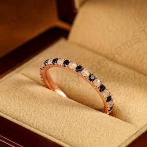 May include: Rose gold ring with alternating black and white gemstones. The ring is set in a brown jewelry box with a beige interior. The ring is a delicate piece of jewelry.