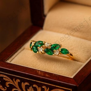 May include: A gold ring with a cluster of emerald-cut green gemstones and small white diamonds. The ring is displayed inside a dark wooden jewelry box with ornate carvings.