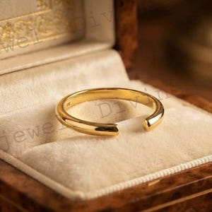 Puede incluir: Un anillo abierto de oro pulido con una banda lisa y redondeada. El anillo se muestra descansando dentro de un joyero de madera forrado con tela de terciopelo color crema, que cuenta con una ranura para sujetar el anillo firmemente.
