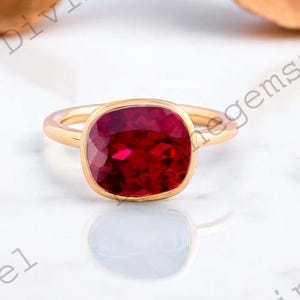 May include: A gold ring featuring a large, square-cut, deep red gemstone. The gemstone is set in a bezel setting, and the band is polished gold. The ring is on a white surface with a blurred background.