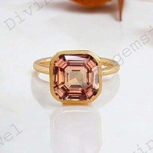 May include: A gold ring with a large, square-cut, peach-coloured gemstone. The gemstone is set in a gold bezel, and the ring band is also gold. The ring is displayed on a white surface with dried leaves in the background.