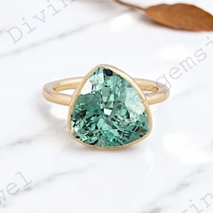Puede incluir: Un anillo de oro con una piedra preciosa verde triangular. El anillo está colocado sobre una superficie de mármol blanco.