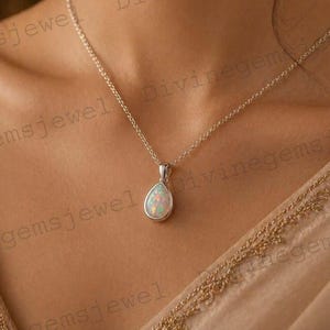 May include: A silver necklace with a pear-shaped opal pendant. The opal has a milky white base with flashes of color. The chain is delicate and hangs on a person's neck. The background is a soft, neutral color.