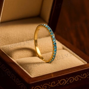 Könnte beinhalten: Ein goldener Ring mit kleinen, runden, blauen Edelsteinen. Der Ring wird in einer hölzernen Schmuckschatulle mit hellbraunem Innenraum präsentiert. Die Schatulle hat eine dunkelbraune Oberfläche mit dekorativen Schnitzereien.