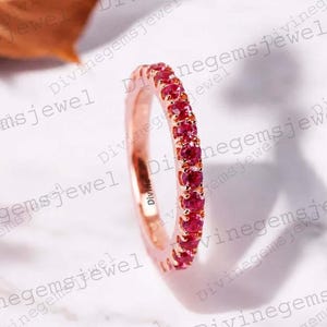 Puede incluir: Una alianza de oro rosa con una hilera continua de pequeñas piedras preciosas redondas de color rojo vibrante, engastadas con garras compartidas. El anillo tiene un acabado metálico pulido y un perfil delgado con piedras uniformes.