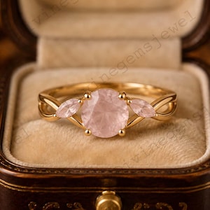May include: A gold ring featuring a large, round, faceted pale pink rose quartz center stone flanked by two smaller, marquise-cut rose quartz stones. The band has a delicate, intertwined design, and the ring is presented in a dark brown box.