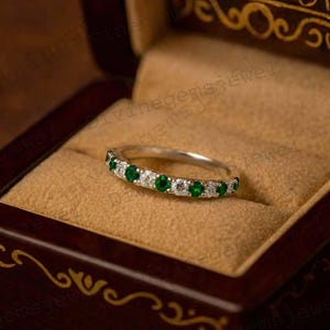 Peut inclure: Une bague en argent ornée d'émeraudes et de diamants alternés, présentée dans un écrin à bijoux en bois foncé avec un intérieur beige. La bague est de style alliance, avec les pierres précieuses rapprochées. L'écrin a des accents décoratifs dorés.