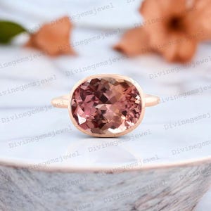 May include: A rose gold ring with a large oval-shaped pink gemstone. The gemstone has a gradient of color from light pink to a darker pink.