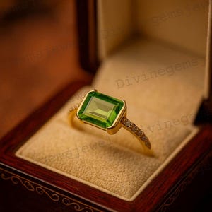 May include: A gold ring with a rectangular, emerald-cut green gemstone. The ring is set in a gold band with small accent stones. The ring is displayed in a wooden jewelry box with a cream-colored interior.