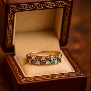 Puede incluir: Un anillo de oro con dos filas de piedras preciosas ovaladas de color verde oscuro y pequeñas piedras preciosas redondas y claras. El anillo se muestra dentro de un joyero marrón oscuro y ornamentado con adornos dorados.
