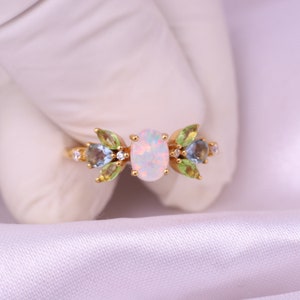 May include: A gold ring featuring a central oval opal gemstone flanked by two pear-shaped blue stones and four marquise-cut green stones. Small clear round accent stones are set between the colored gems, creating a floral-inspired arrangement.