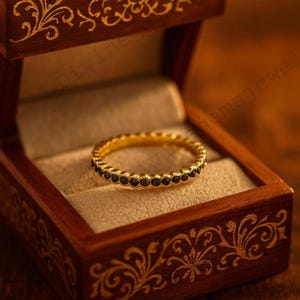 May include: A gold ring with a band of small, black gemstones. The ring is displayed inside a wooden jewelry box with ornate gold detailing. The box is open, revealing the ring against a beige interior.