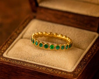 May include: A gold ring with a series of round, vibrant green gemstones set around the band. The ring is displayed inside a wooden jewelry box with a tan interior, creating a luxurious presentation. The ring's design is simple yet elegant.