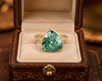 May include: A gold ring with a large, triangular, teal-colored gemstone. The ring is set in a gold band and is displayed in a brown, velvet-lined jewelry box. The box is made of dark wood and has a decorative clasp.