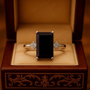 May include: A silver ring with a large, rectangular black gemstone as the centerpiece. Small, diamond-studded square accents flank the main stone. The ring is presented in a dark wooden jewelry box with a light tan interior.