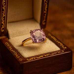 Può includere: Un anello d'oro con una grande pietra preziosa rosa chiaro taglio cuscino. L'anello è presentato in un cofanetto di legno scuro con finiture dorate. La gemma è il punto focale, catturando la luce e riflettendo una morbida lucentezza.