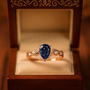 Op de afbeelding: Rosegouden ring met een peervormige, diepblauwe edelsteen. De band heeft een delicaat, bladvormig ontwerp met kleine, heldere accentstenen. De ring wordt gepresenteerd in een houten sieradendoosje.