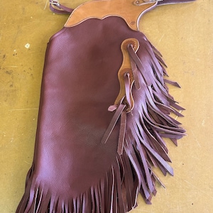 May include: A burgundy leather chaps with a light brown leather yoke and fringe. The chaps have a buckle closure and decorative leather accents. The fringe is long and hangs down the side of the chaps.