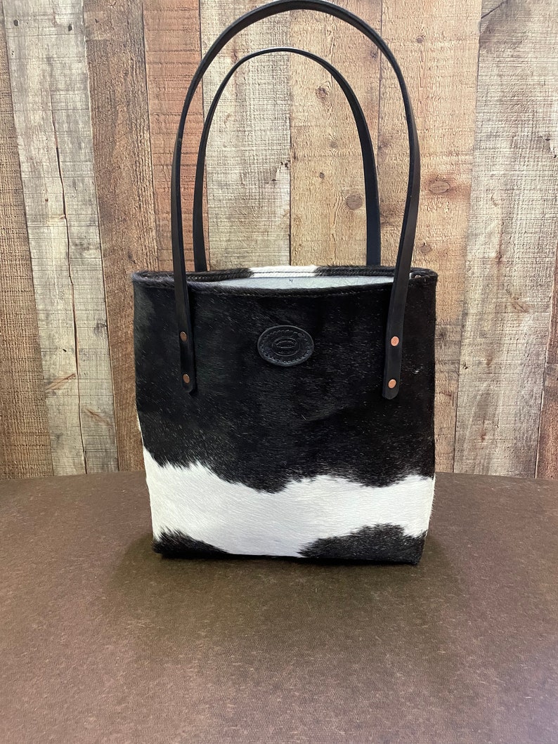 Pu&ograve; includere: Borsa tote in pelle di mucca bianca e nera con manici in pelle nera e un accento in pelle marrone. La borsa ha un design semplice ed &egrave; perfetta per l'uso quotidiano.