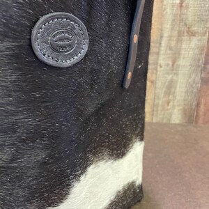 Pu&ograve; includere: Borsa tote in pelle di mucca bianca e nera con manico in pelle e un cerchio in pelle circolare che dice "Artisan Made Leather Goods".