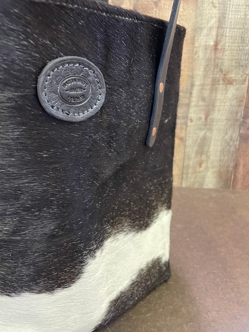 Pu&ograve; includere: Borsa tote in pelle di mucca bianca e nera con manico in pelle e un cerchio in pelle circolare che dice "Artisan Made Leather Goods".