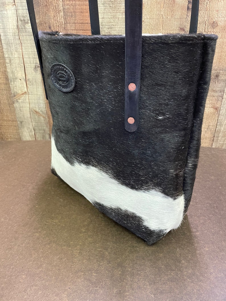 Pu&ograve; includere: Borsa tote in pelle di mucca bianca e nera con cinturini in pelle nera e una toppa in pelle marrone. La borsa ha una chiusura a zip ed &egrave; realizzata con materiali di alta qualit&agrave;.