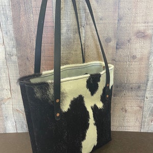Pu&ograve; includere: Una borsa tote in pelle di mucca bianca e nera con cinturini in pelle nera e rivetti in rame. La borsa &egrave; mostrata su uno sfondo in legno.
