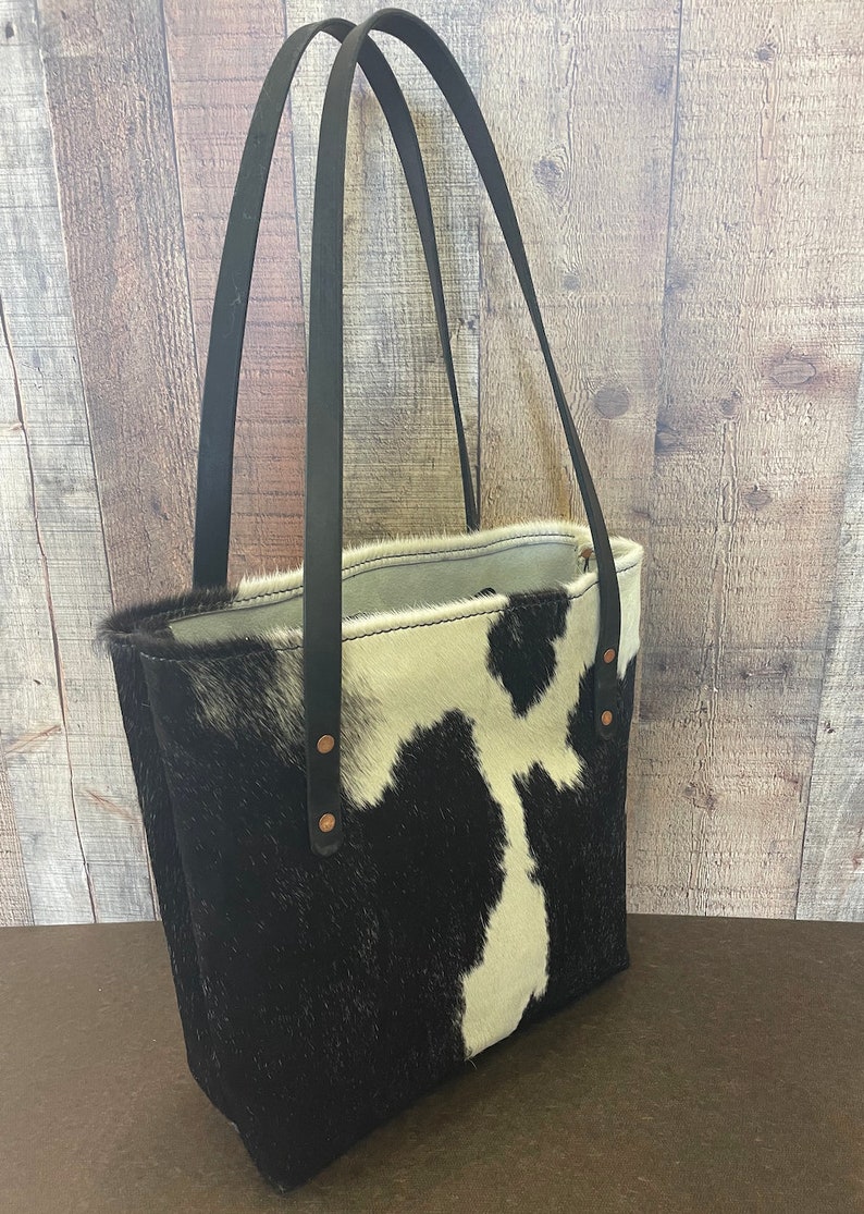 Pu&ograve; includere: Una borsa tote in pelle di mucca bianca e nera con cinturini in pelle nera e rivetti in rame. La borsa &egrave; mostrata su uno sfondo in legno.