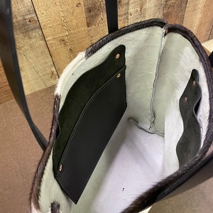 Pu&ograve; includere: Una borsa tote in pelle di mucca bianca e nera con interno in pelle nera e una tasca in pelle nera. La borsa ha una tracolla in pelle nera.