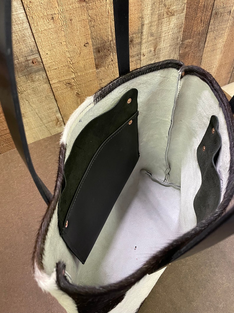 Pu&ograve; includere: Una borsa tote in pelle di mucca bianca e nera con interno in pelle nera e una tasca in pelle nera. La borsa ha una tracolla in pelle nera.
