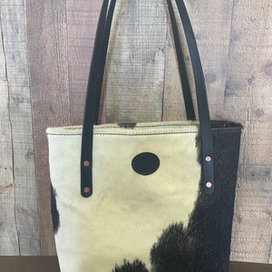 Pu&ograve; includere: Una borsa tote in pelle di mucca bianca e nera con manici in pelle nera e rivetti in rame. La borsa ha una toppa ovale in pelle nera sul davanti.
